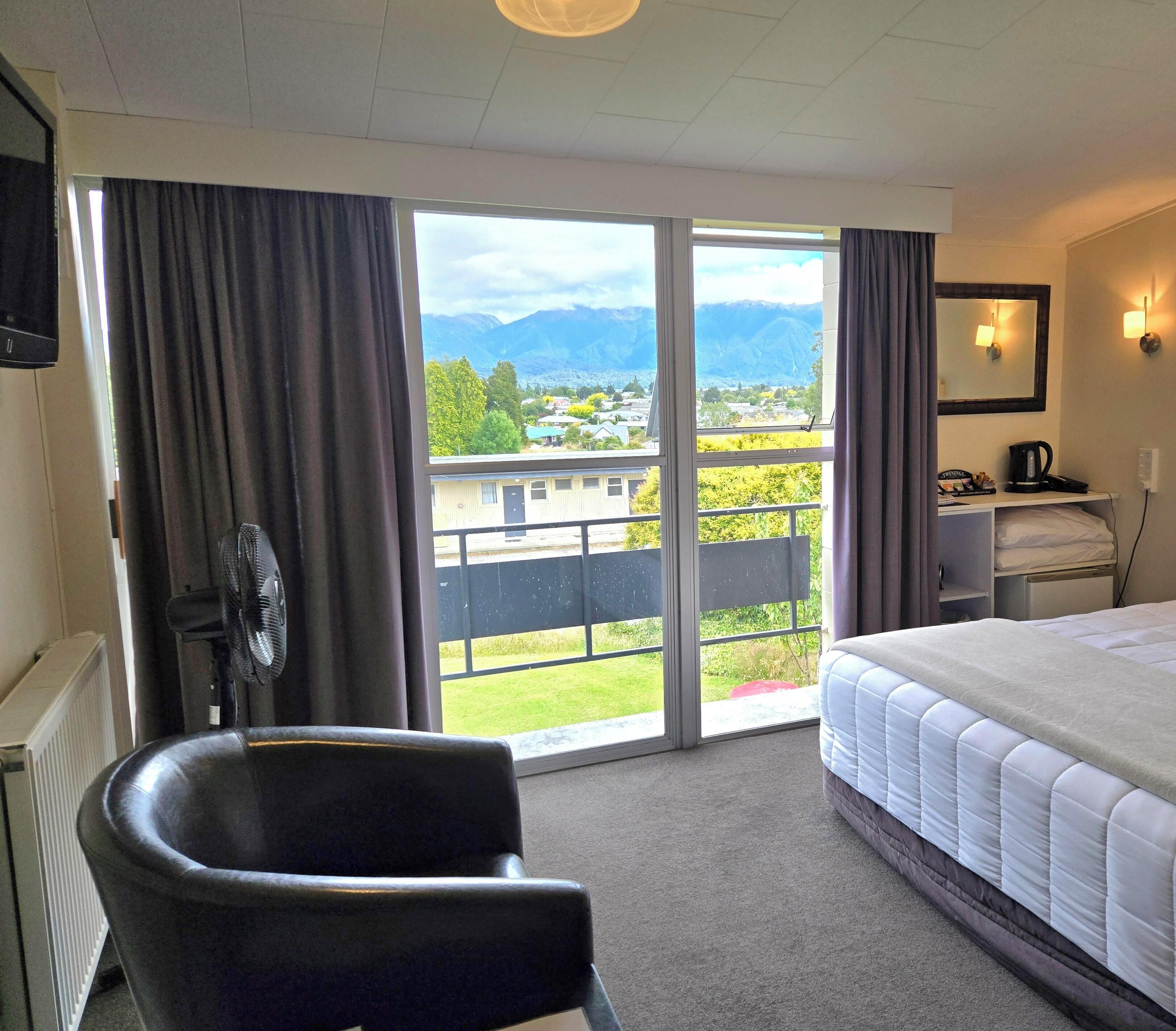 A hotel room with a bed, a chair, a desk with a lamp, and a view of a town and mountains through the window.