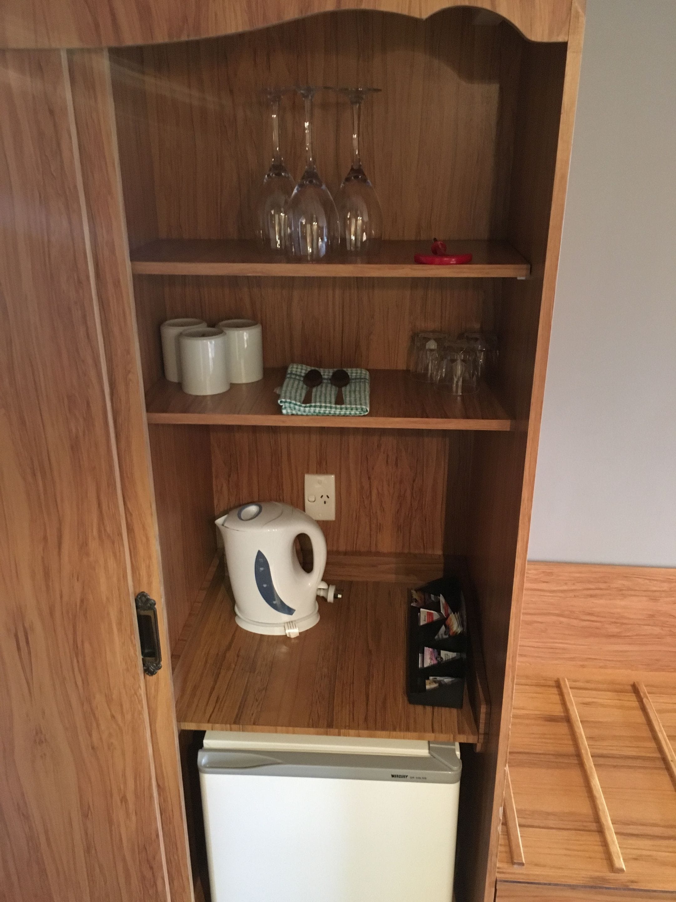 A wooden cabinet with shelves containing wine glasses, a kettle, and a checkered cloth.