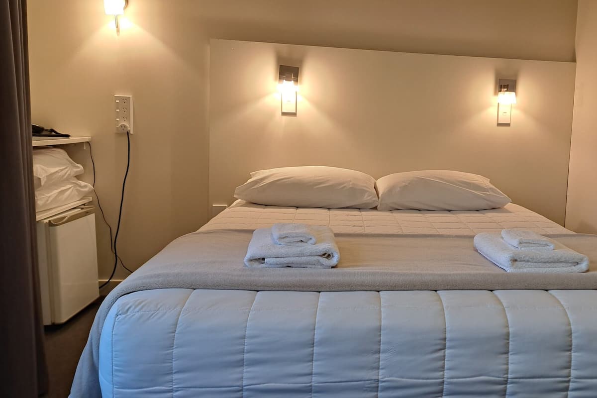 Una cama bien hecha con dos almohadas y dos toallas dobladas, una mesita de noche con una lámpara y un enchufe en la pared.
