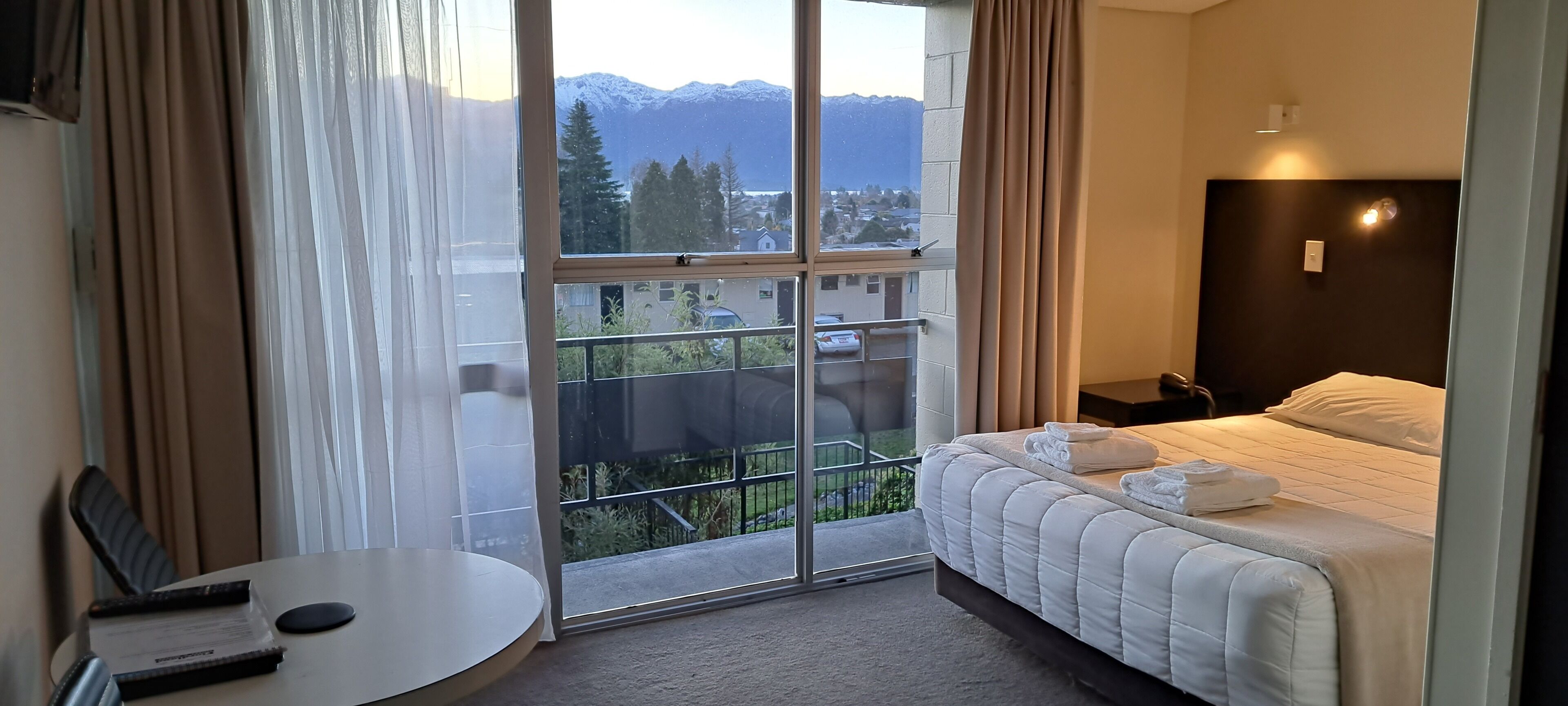 A hotel room with a bed, a small table, and a window with a view of mountains and a city.