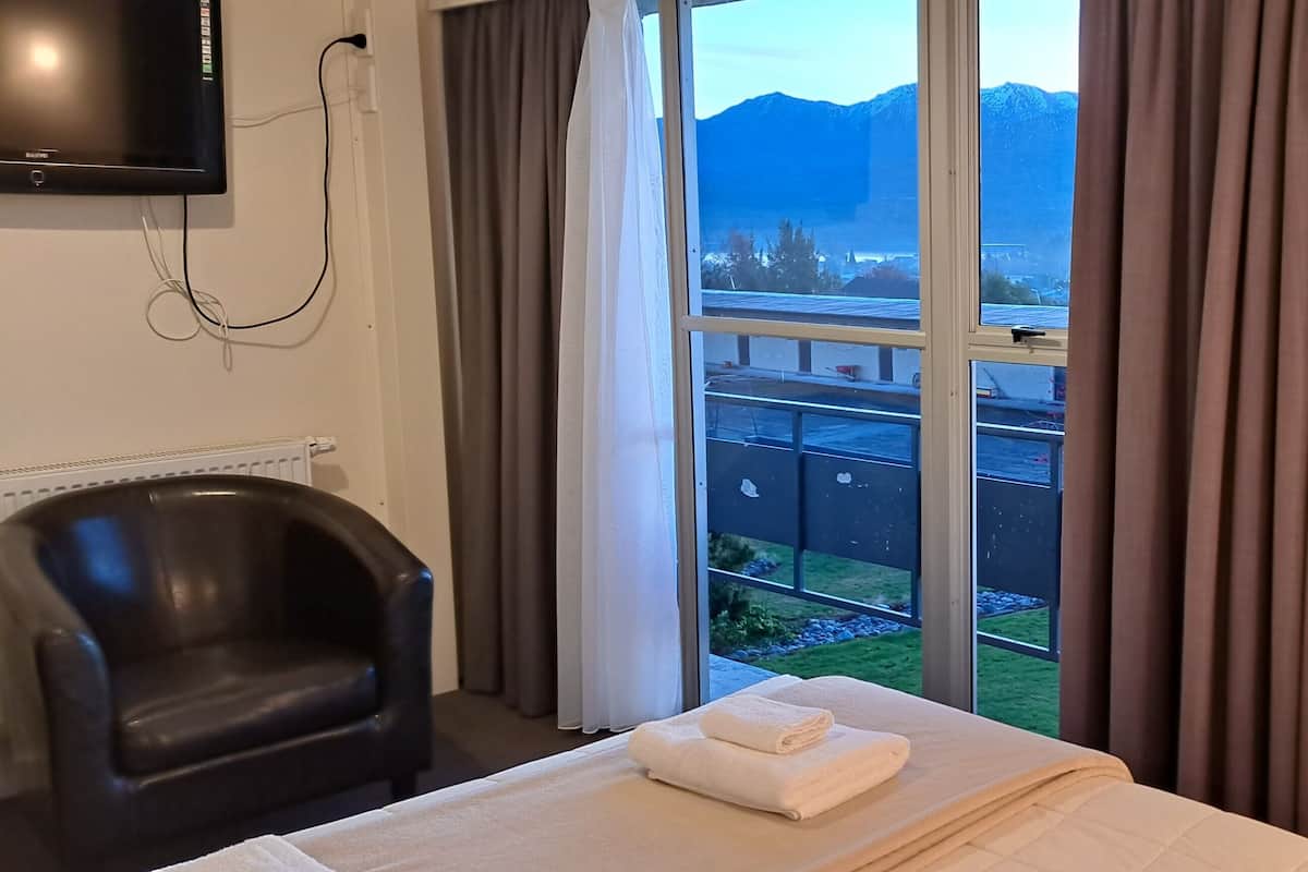 Habitación de hotel con cama, una silla, televisión, ventana con cortinas y estante con artículos de aseo.