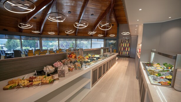 A buffet area with various food items, wooden ceiling, and large windows offering a view of the outdoors. - Distinction Luxmore Hotel (Te Anau)