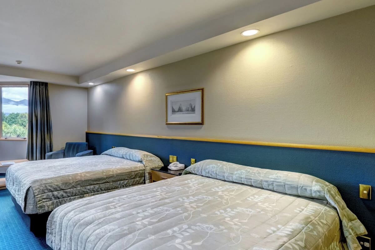 A hotel room with two beds, a desk, a chair, and a large window offering a mountain view.