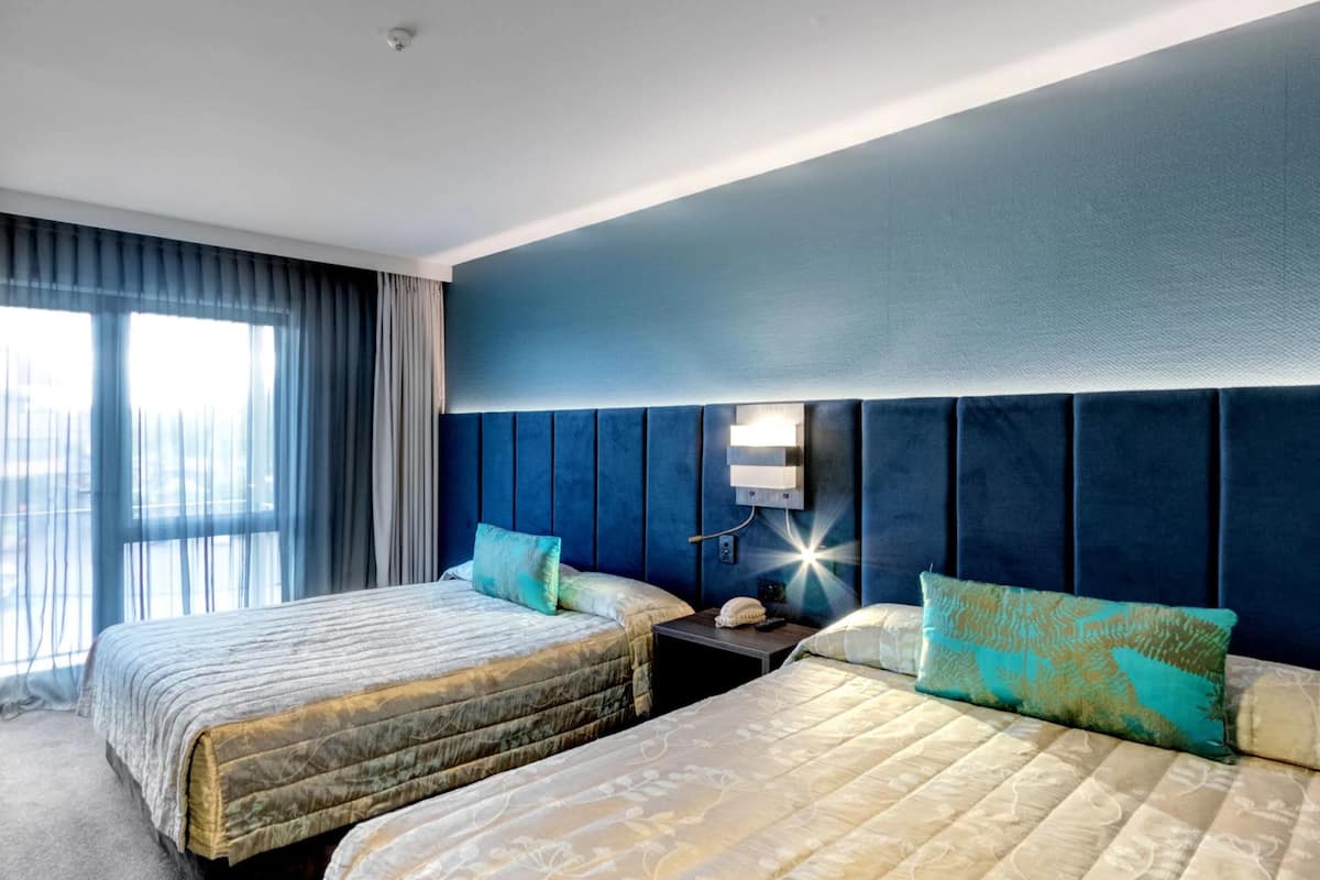 A hotel room with two beds, a nightstand, a wall-mounted light, and a large window with curtains.