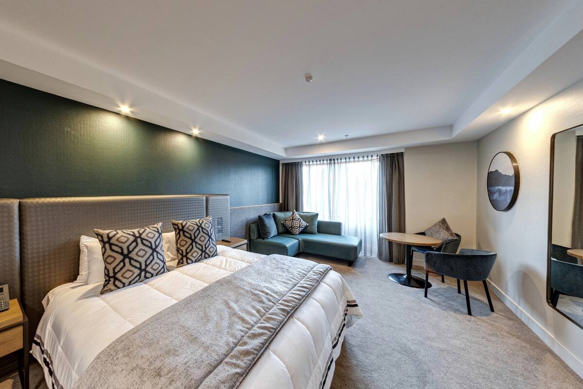 A modern hotel room with a large bed, a sofa, a small table, and a mirror.