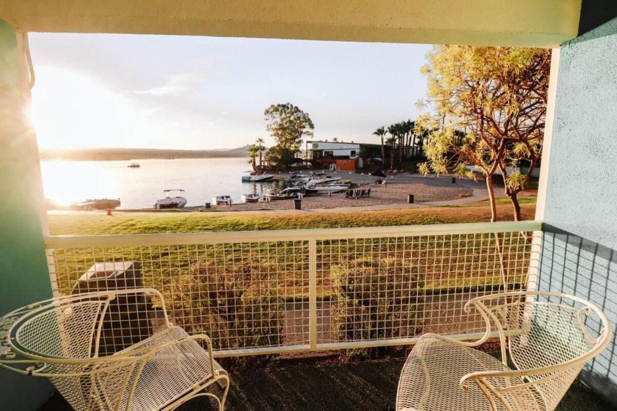 Un balcon avec des fauteuils en osier, une vue sur une plage avec des bateaux, et un bâtiment en arrière-plan.