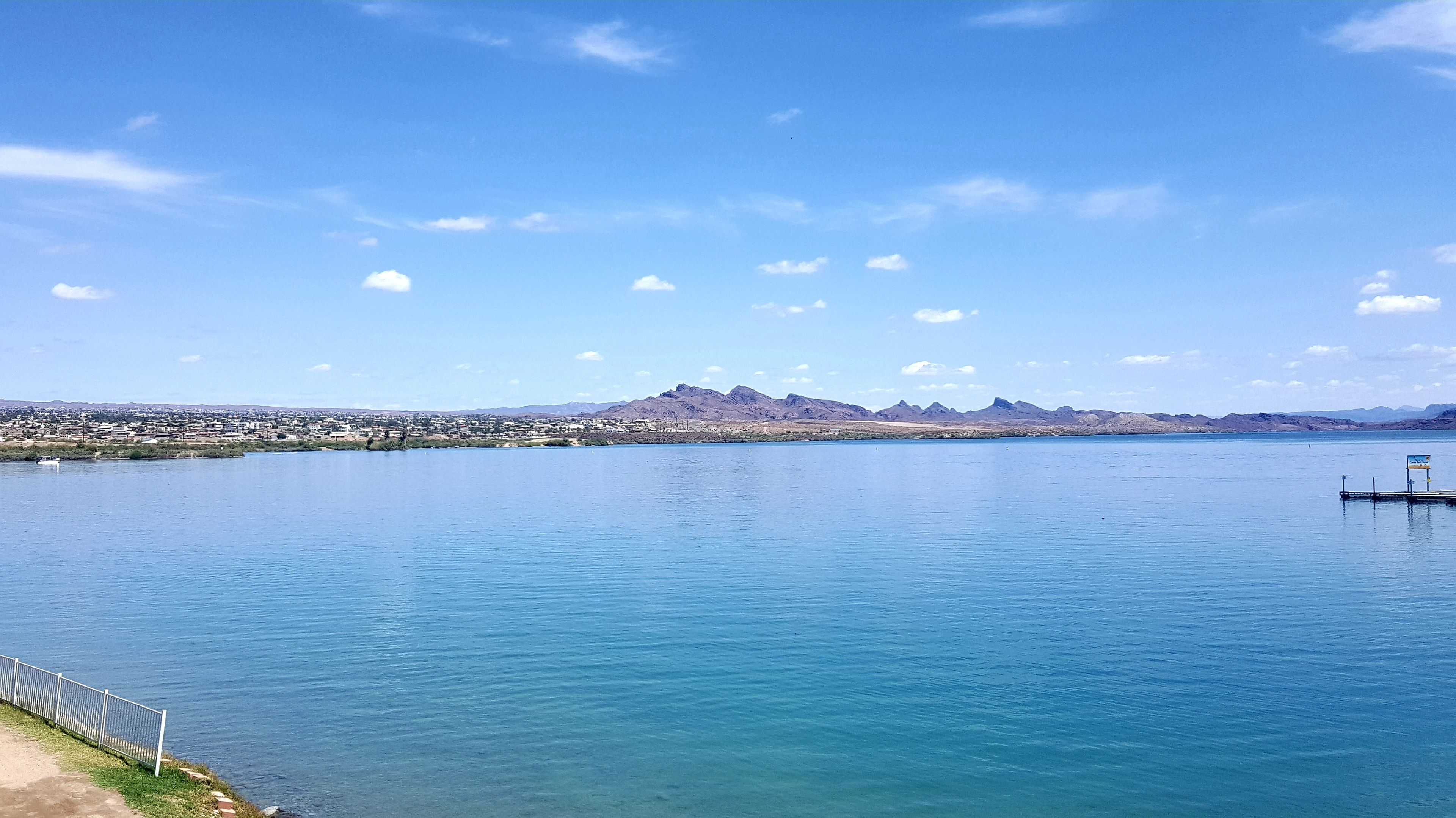 Un lac paisible, avec un quai et un panneau, entouré de montagnes et d’une ville qui se devine au loin.