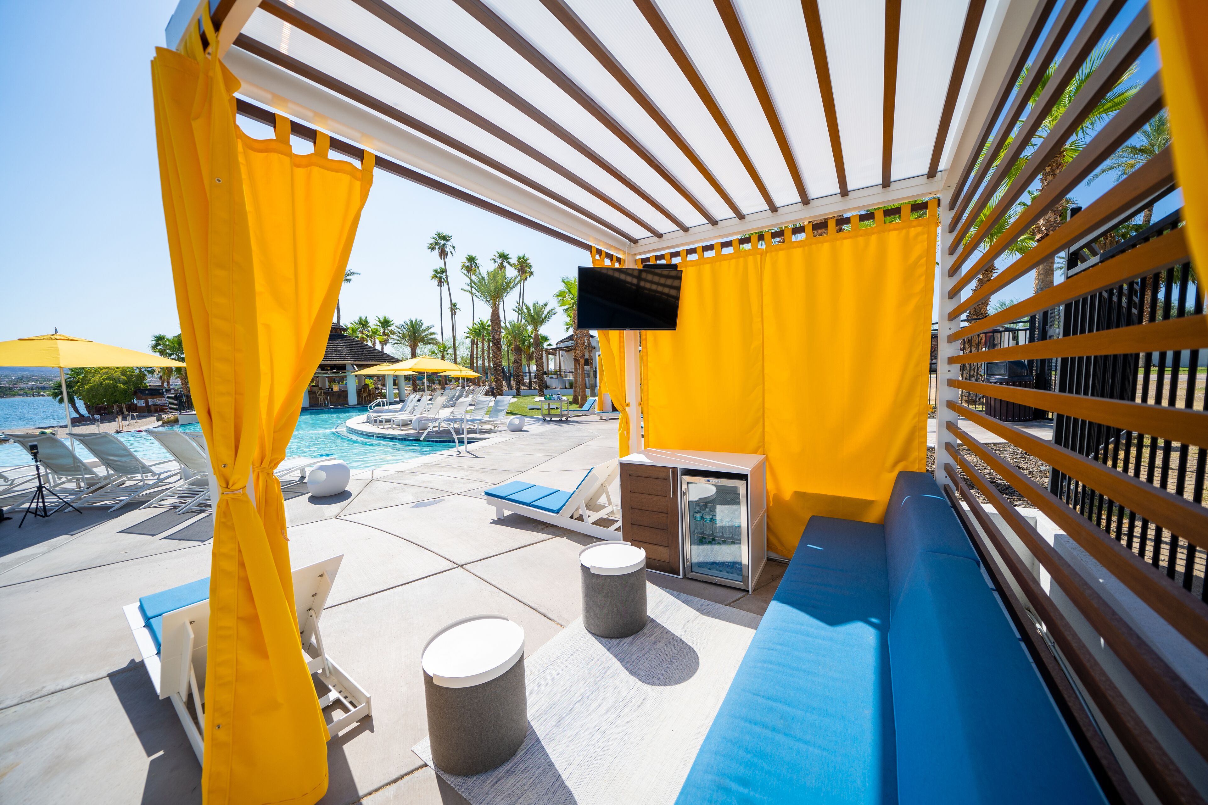 A poolside area with yellow curtains, lounge chairs, a TV, and a view of the pool and palm trees.