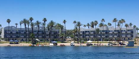 A beachfront hotel with multiple floors, palm trees, and a sandy beach area with umbrellas and boats.
