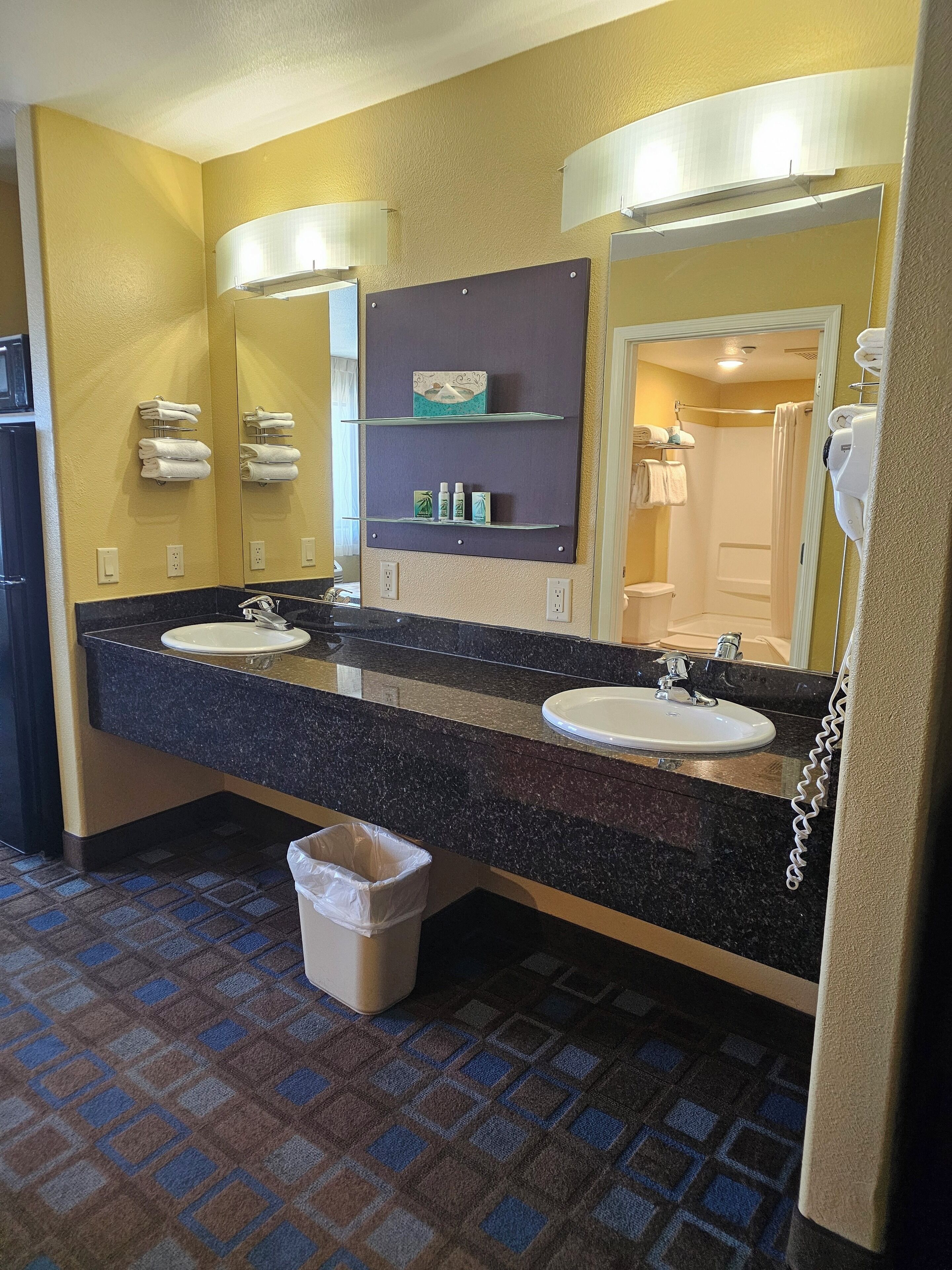 A hotel bathroom with two sinks, a mirror, towel racks, and a trash bin.