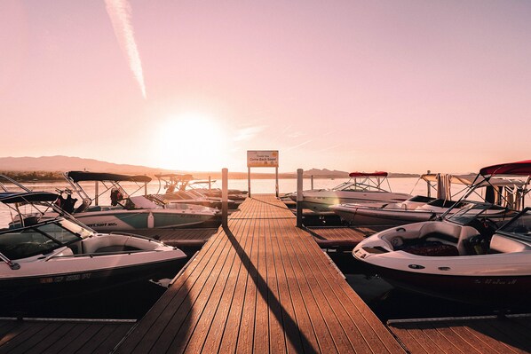 Un muelle de madera con barcos amarrados a ambos lados, un cartel que dice '¡Gracias, vuelva pronto!' y un atardecer en el fondo.