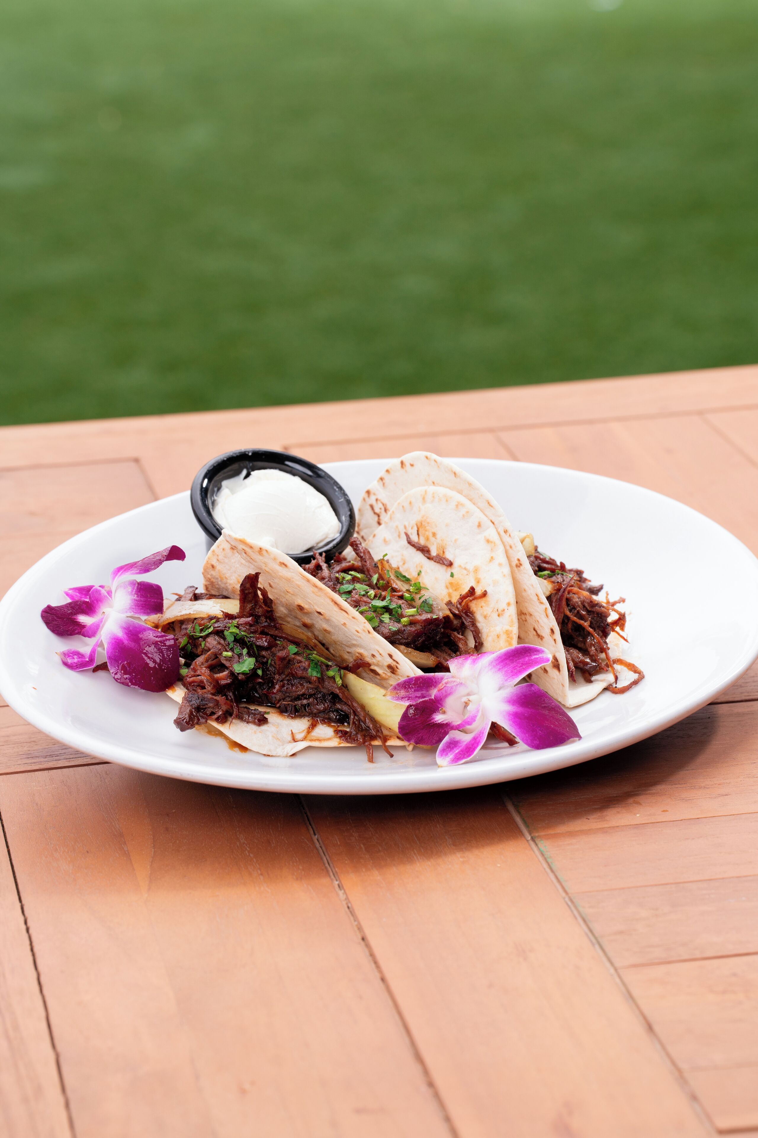 Un plato de tacos acompañado de crema agria y flores moradas decorativas sobre una mesa de madera.