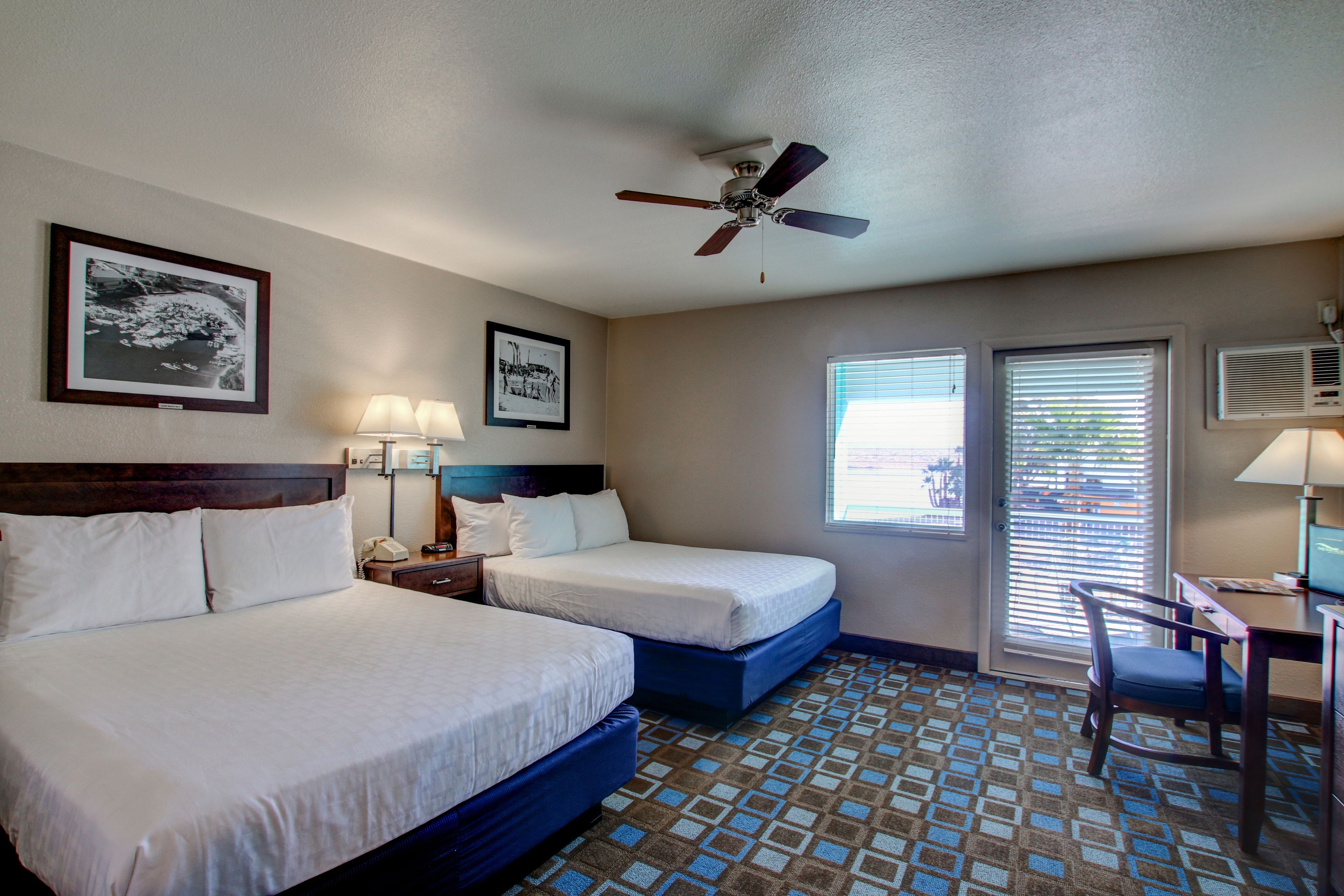 A hotel room with two beds, a ceiling fan, a desk with a chair, and two windows with blinds.
