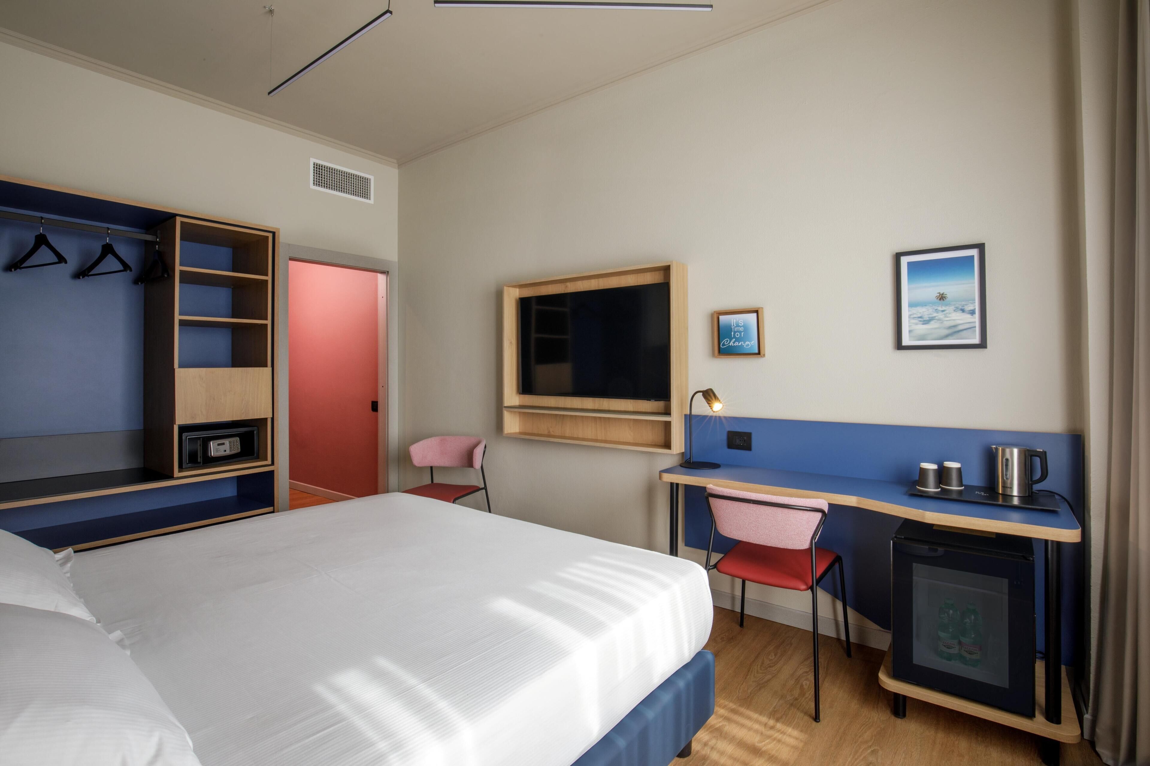 A hotel room with a bed, a desk, a chair, a wardrobe, a TV, and a small refrigerator.