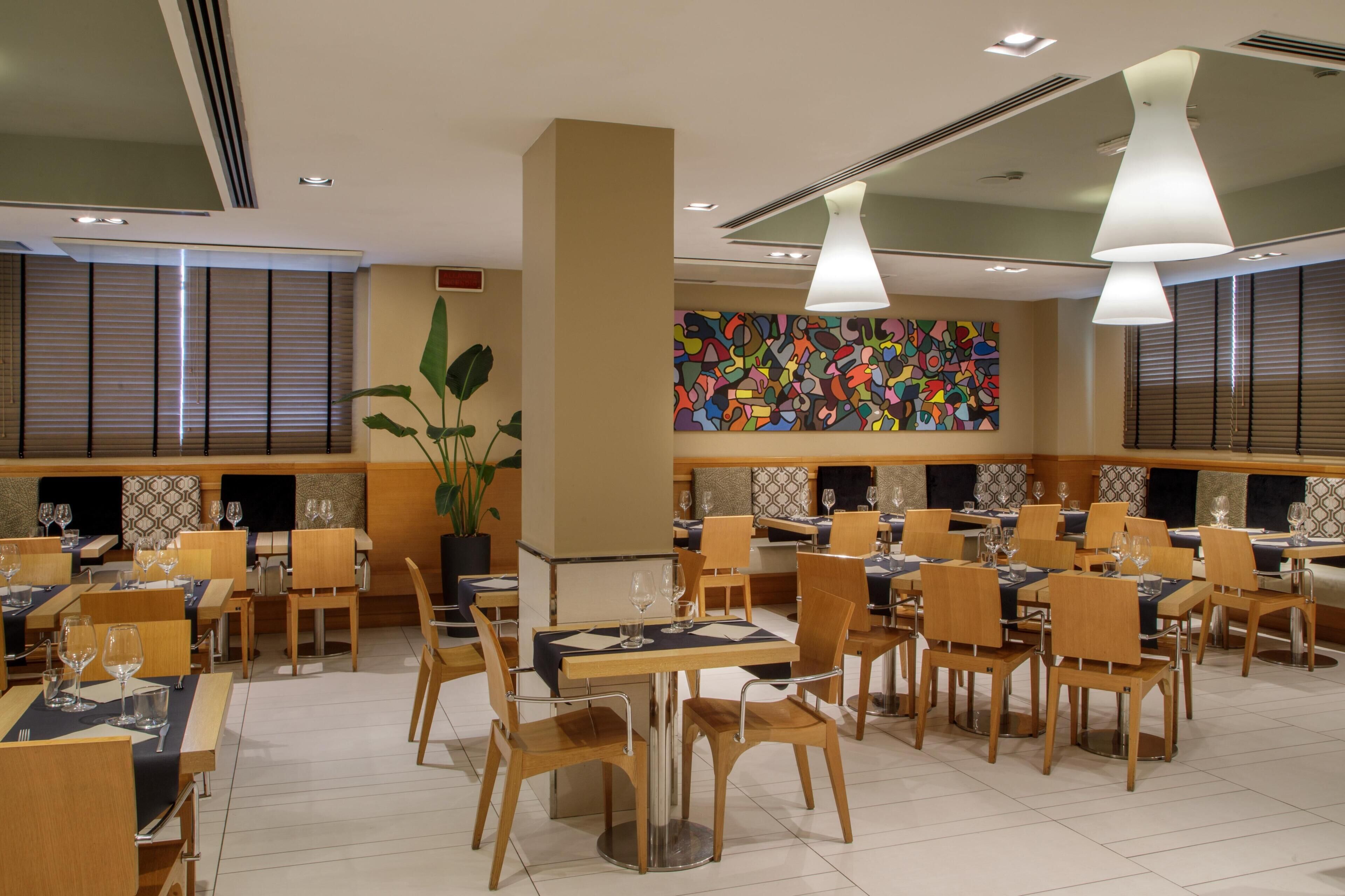 Un restaurant avec plusieurs tables et chaises, une œuvre d’art abstraite et colorée sur le mur, et une plante en pot.