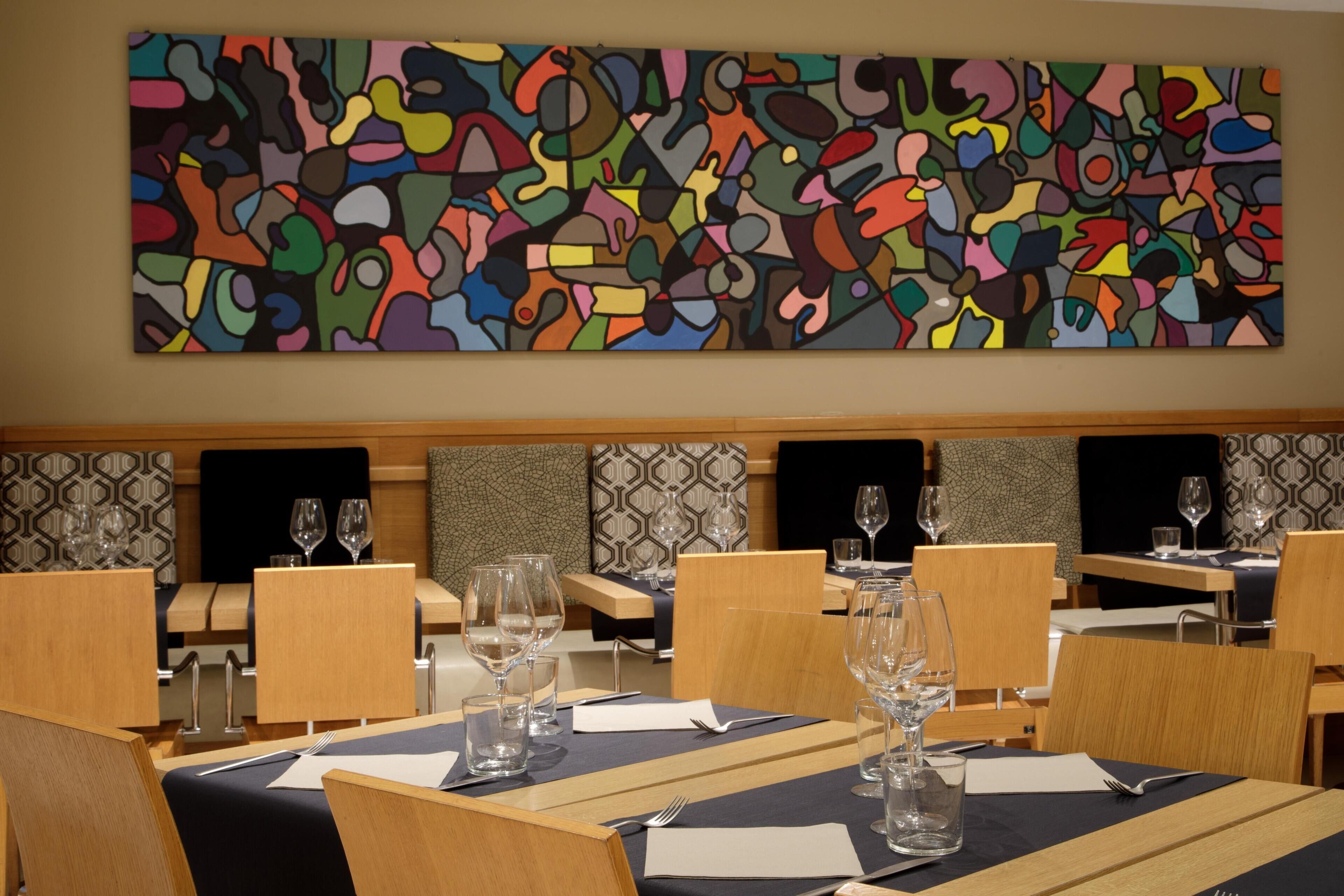 Een eetgedeelte met tafels voor twee personen, elk voorzien van wijnglazen en bestek. Een kleurrijk abstract schilderij bevindt zich boven de tafels aan de muur.
