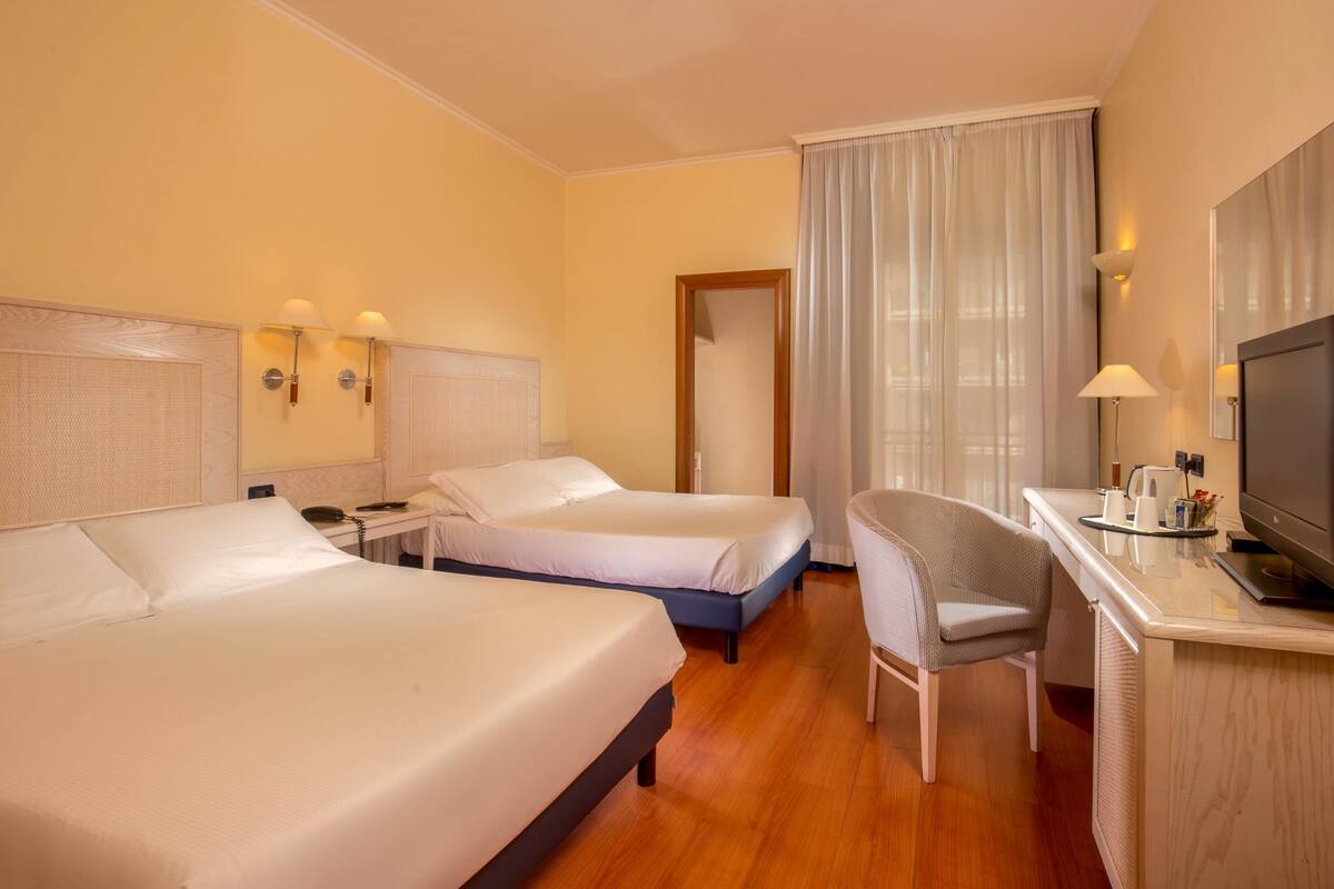 Hotellihuone, jossa on kaksi sänkyä, työpöytä tuolilla, televisio ja suuri ikkuna, jossa on verhot.