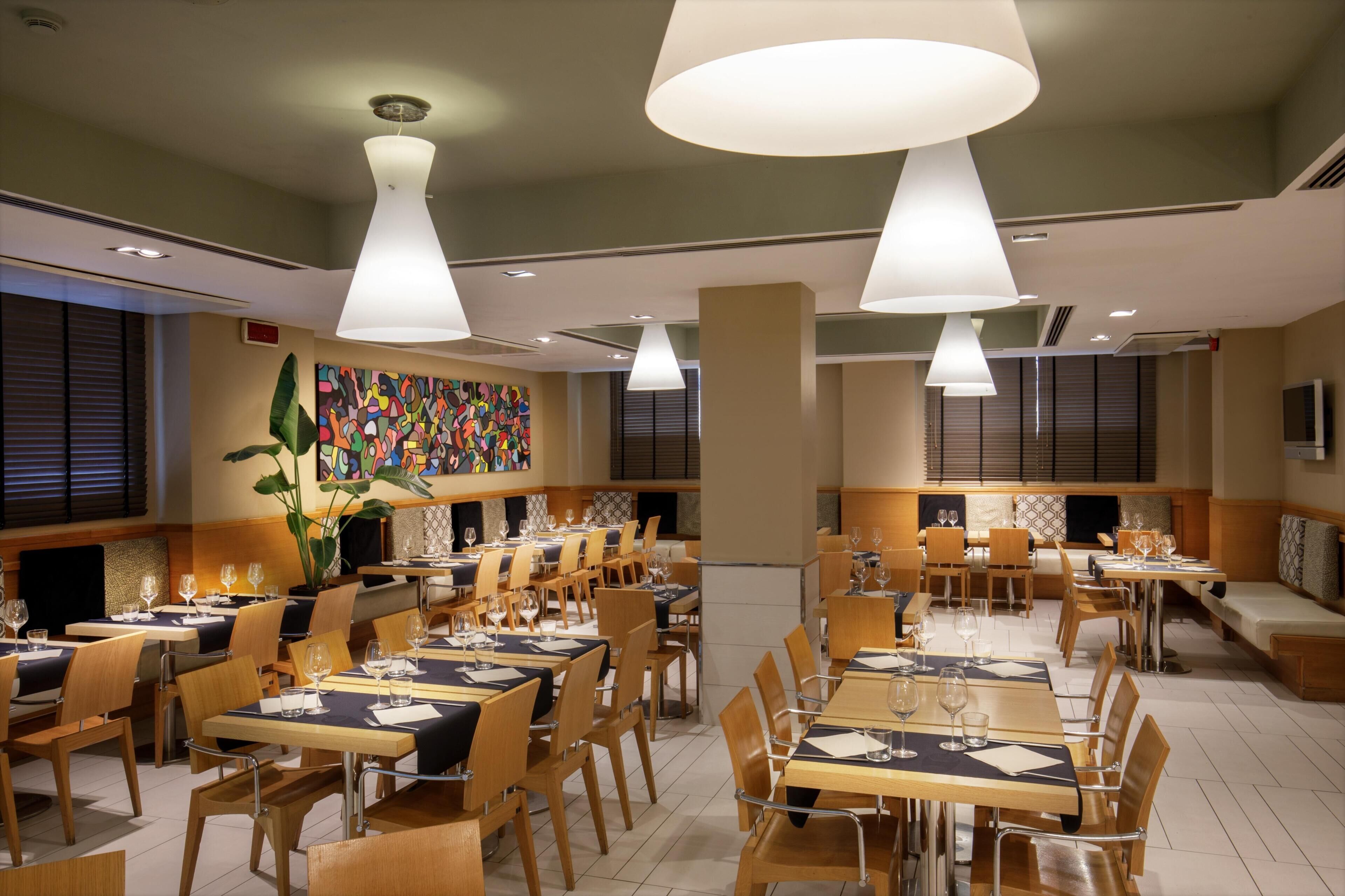 Un restaurante con varias mesas, sillas, iluminación colgante y un colorido cuadro abstracto en la pared.