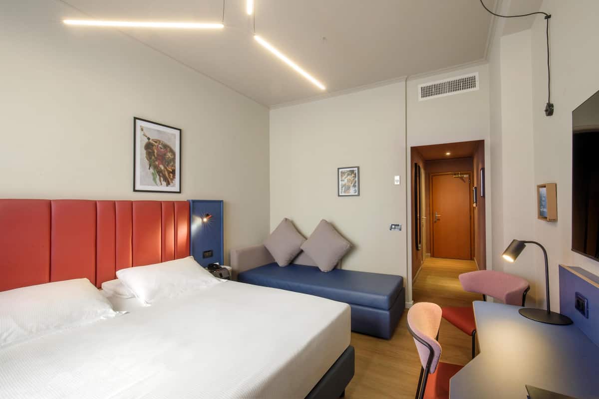 Hotellihuoneessa on sänky, sohva, työpöytä ja tuoli. Huoneesta on ovi toiseen huoneeseen.
