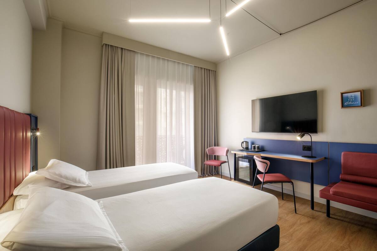Hotellihuone, jossa on kaksi sänkyä, työpöytä tuolilla, televisio ja ikkuna, jossa on verhot.