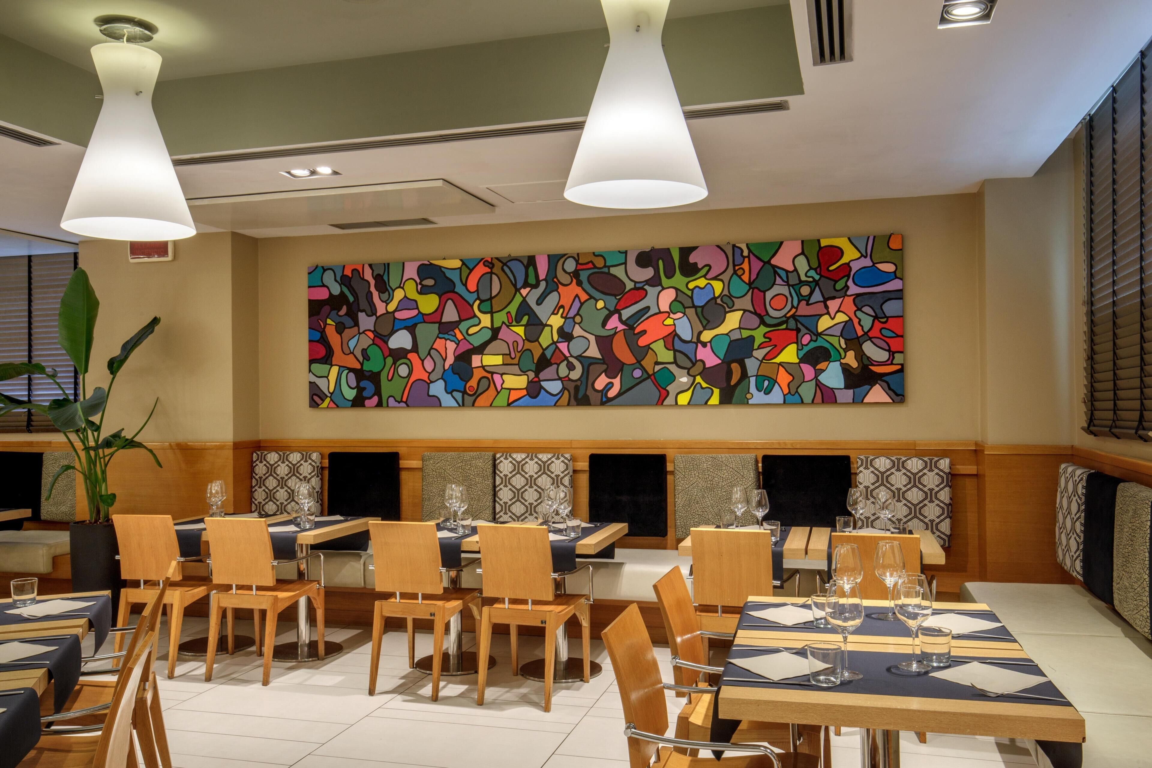 Un restaurante con varias mesas y sillas, un cuadro abstracto de colores en la pared, e iluminación colgante.