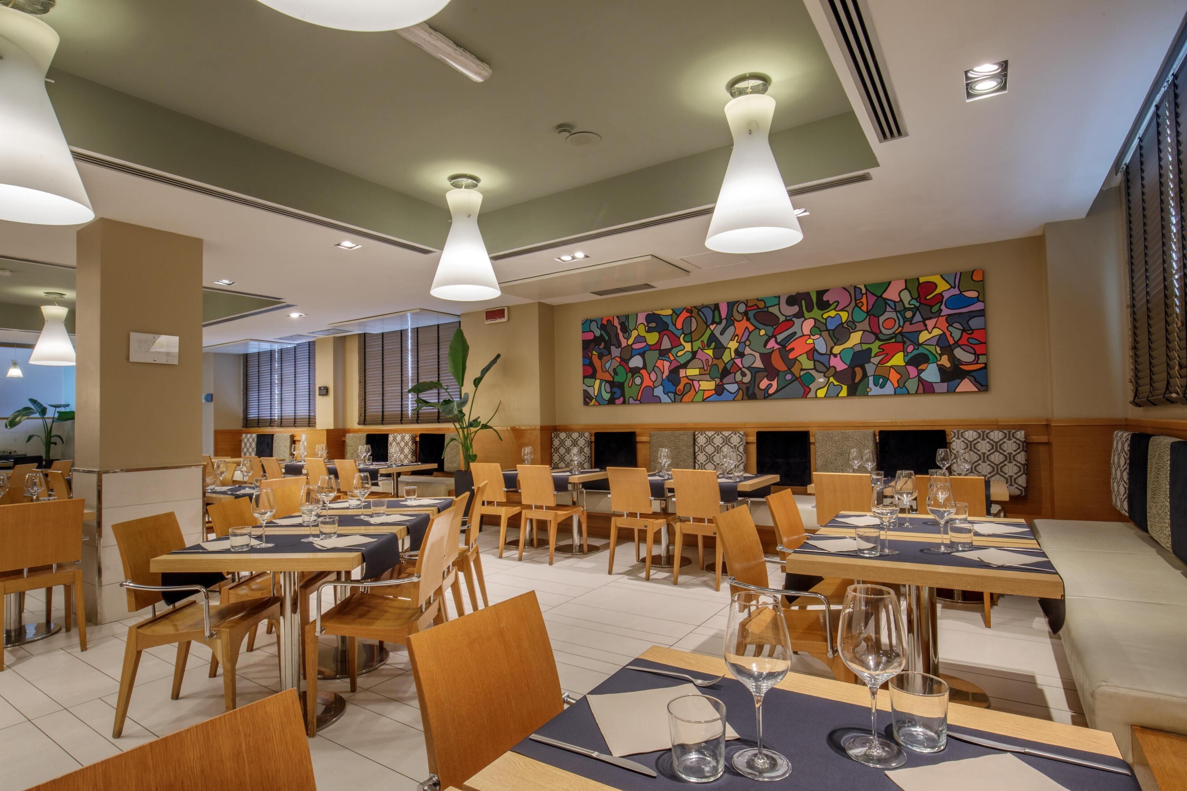 A restaurant with multiple dining tables, chairs, pendant lighting, and a colorful abstract wall art.