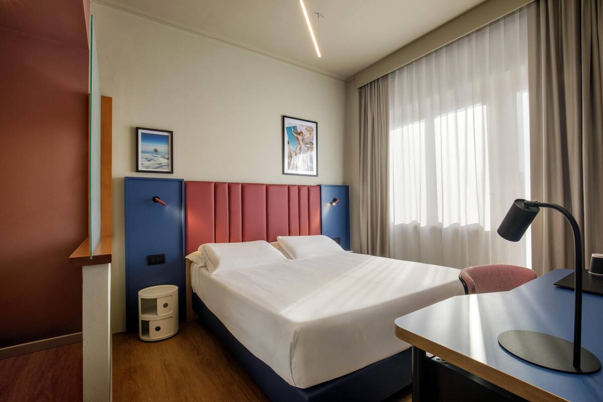 Hotellihuone, jossa on sänky, työpöytä, tuoli, yöpöytä, lamppu ja kaksi kehystettyä kuvaa seinällä.