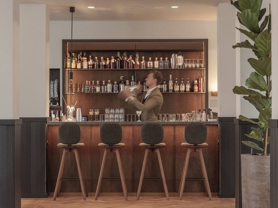 Una zona de bar con un barman preparando bebidas, taburetes de madera y una estantería con varias botellas.