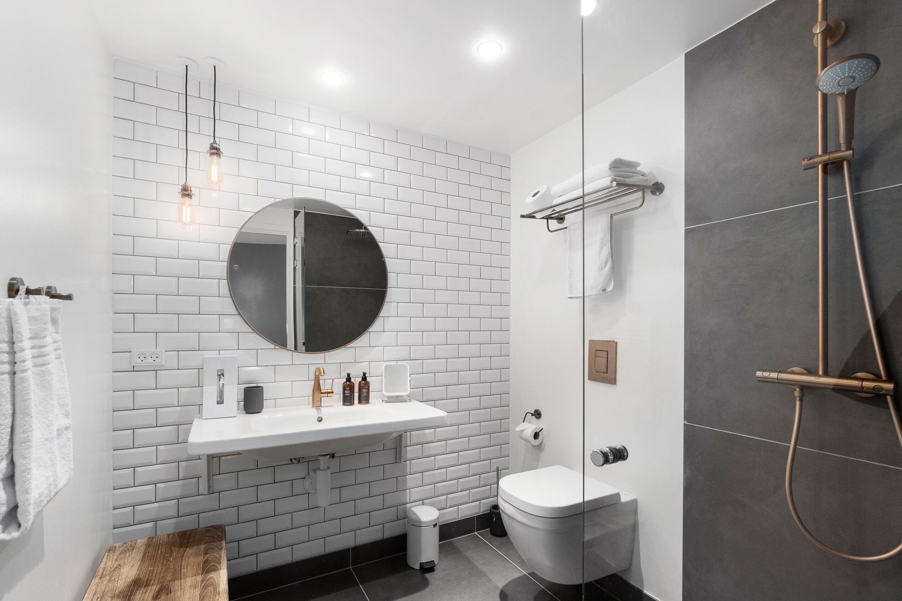 A modern bathroom with a white tiled wall, a round mirror, a white sink, a toilet, and a shower area.