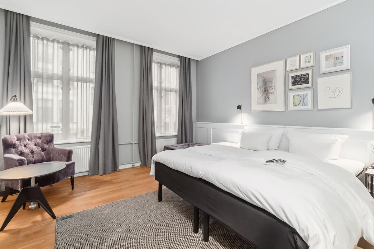 A modern bedroom with a large bed, a round table, and a chair. There are curtains on the windows, and artwork on the wall.