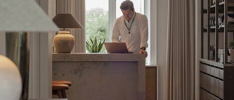 A person at a desk with a laptop, a vase, and a plant in a room with a large window and wooden flooring.