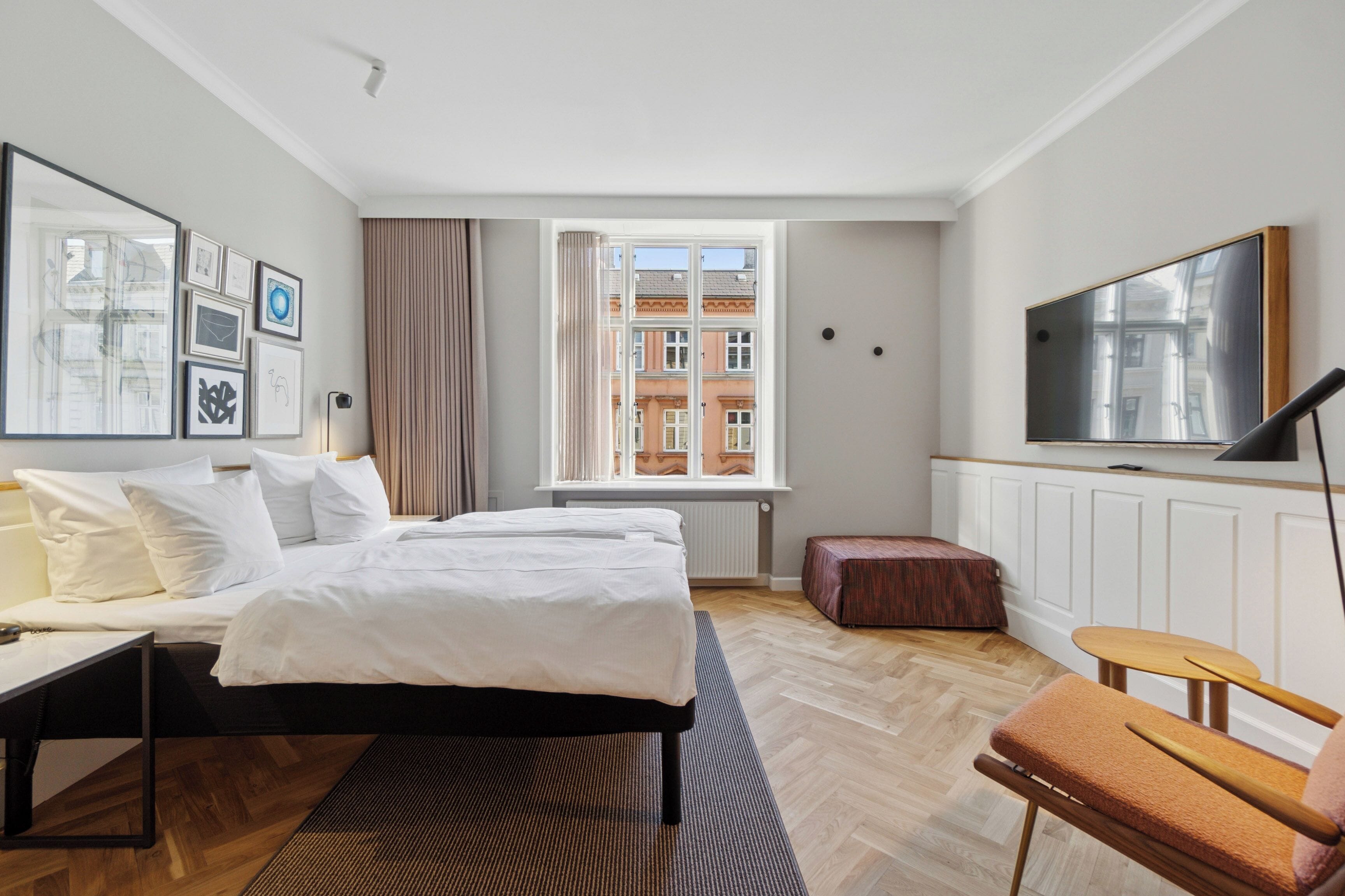 A hotel room with a bed, a desk, a chair, a TV, and a window with a view of buildings.