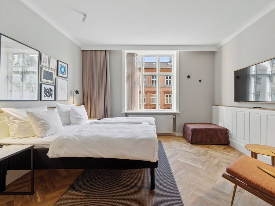 A hotel room with a bed, a desk, a chair, a TV, and a window with a view of buildings.
