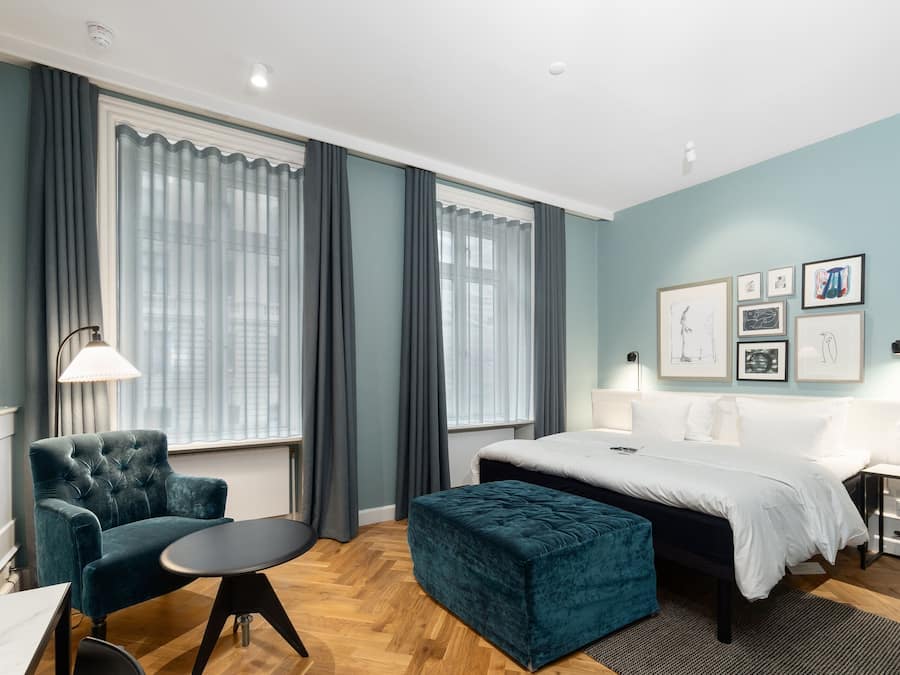 Una habitación de hotel moderna con una cama grande, un sillón capitonado, una mesita pequeña y un banco.