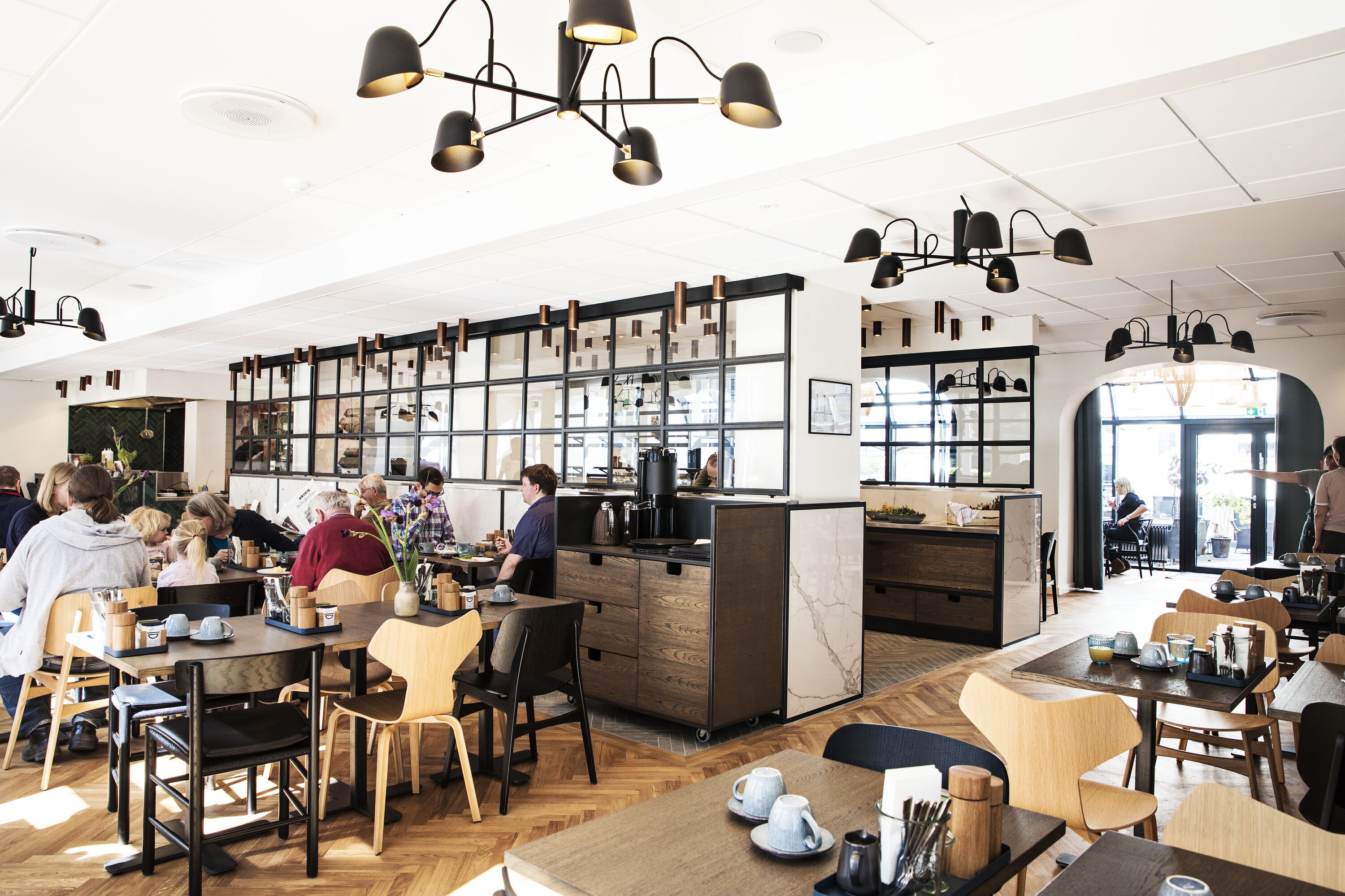 Un café moderno con mesas y sillas de madera, una barra central y lámparas colgantes.