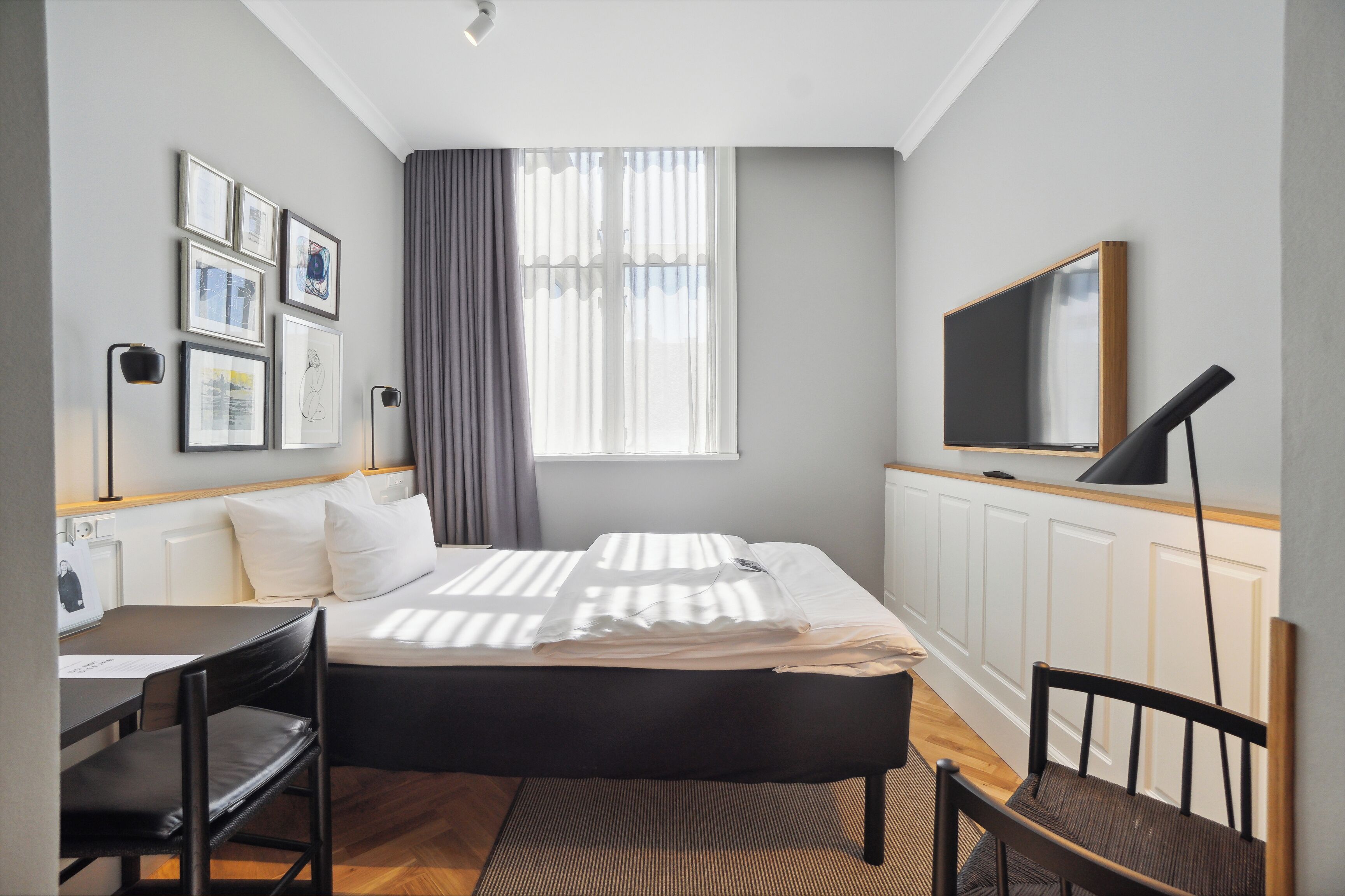 Habitación de hotel pequeña con cama, escritorio, silla, televisor y ventana con cortinas.