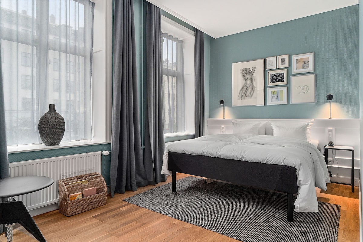 A bedroom with a bed, a window with curtains, a radiator, a bedside table, a lamp, and framed pictures on the wall.