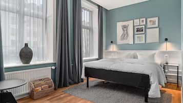 A bedroom with a bed, a window with curtains, a radiator, a bedside table, a lamp, and framed pictures on the wall.