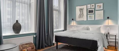 A bedroom with a bed, a window with curtains, a radiator, a bedside table, a lamp, and framed pictures on the wall.