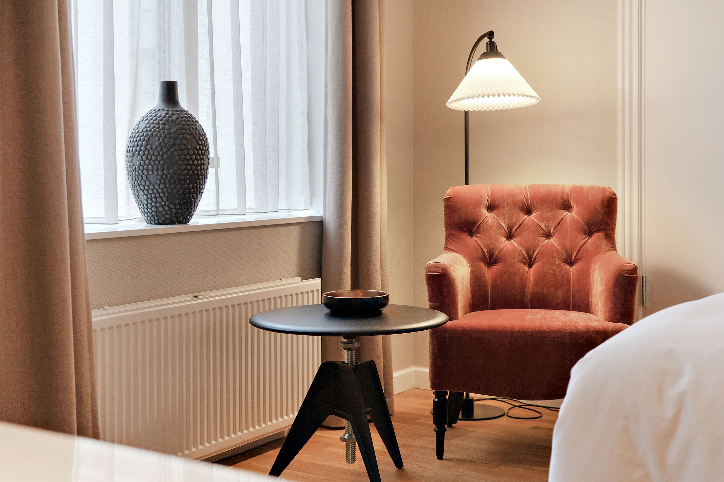 Ein Zimmer mit Fenster, Heizkörper, rundem Tisch mit Schale, Ohrensessel und Stehlampe.