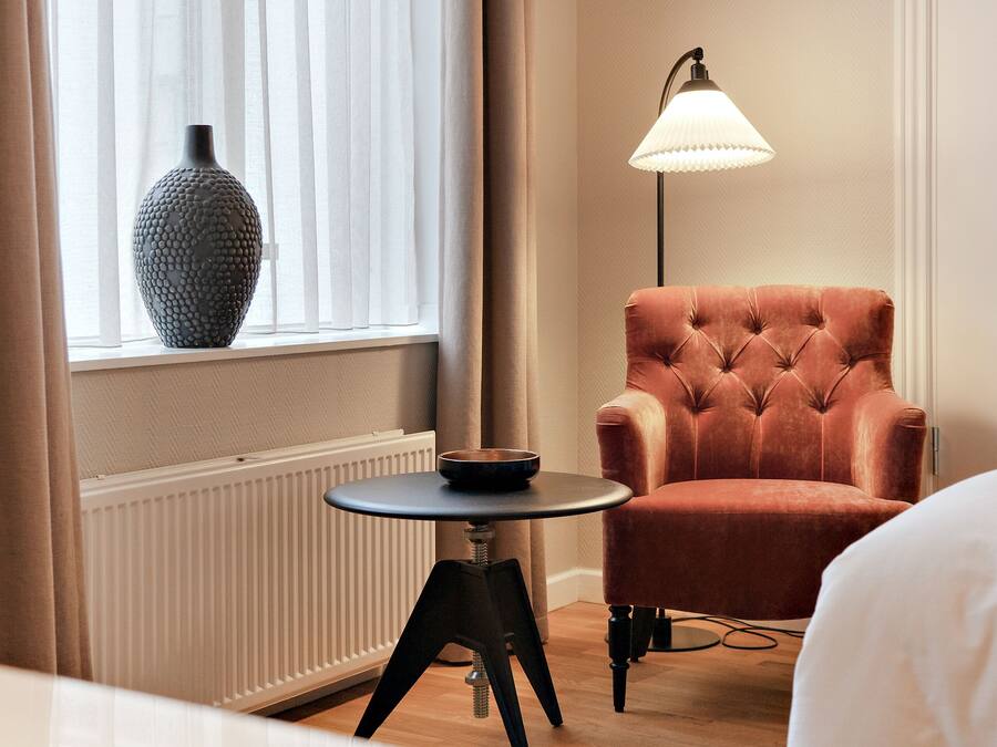 Ett rum med ett fönster, ett element, ett runt bord med en skål på, en stoppad fåtölj och en golvlampa.