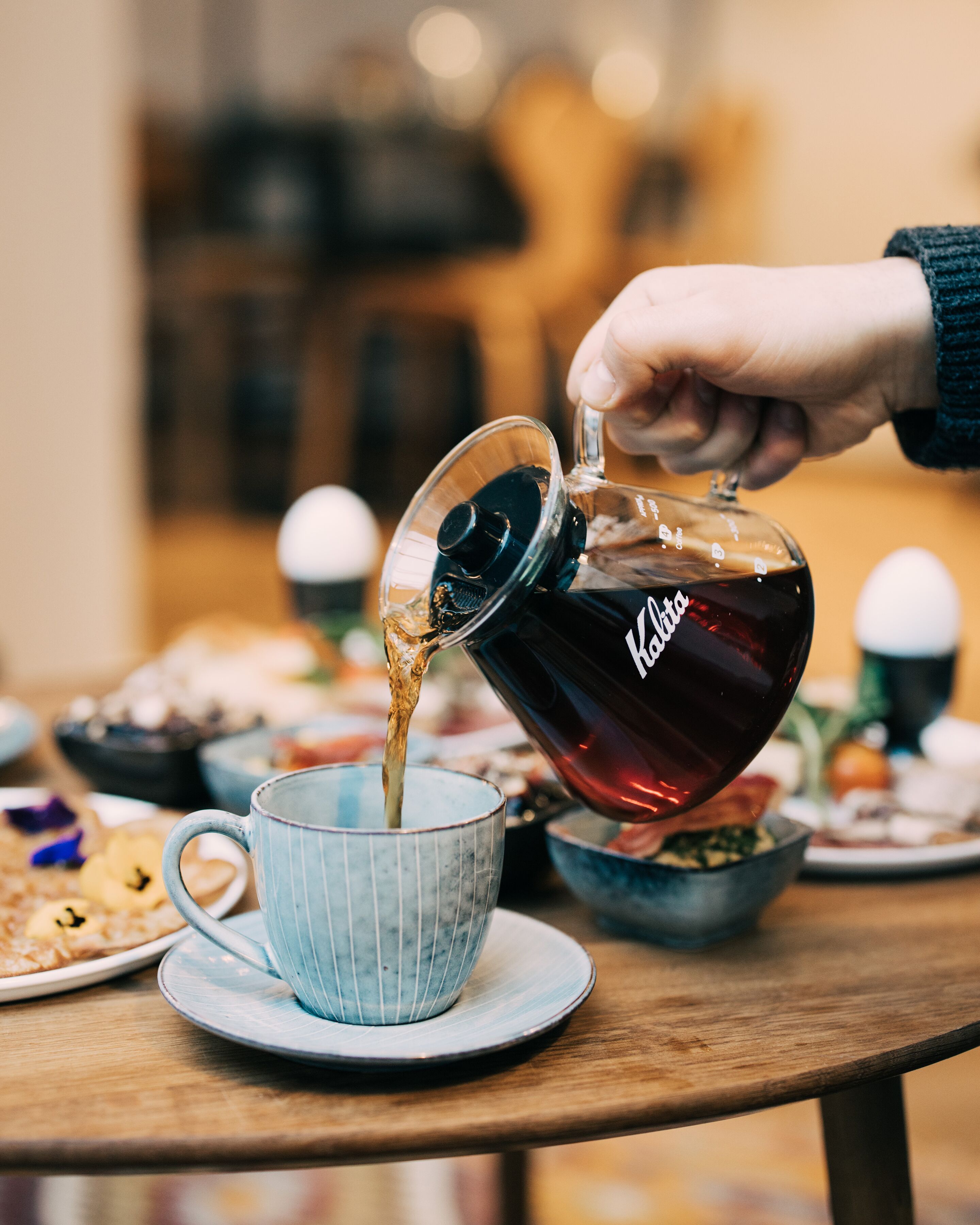 A hand pouring tea from a glass teapot into a ceramic cup on a wooden table.