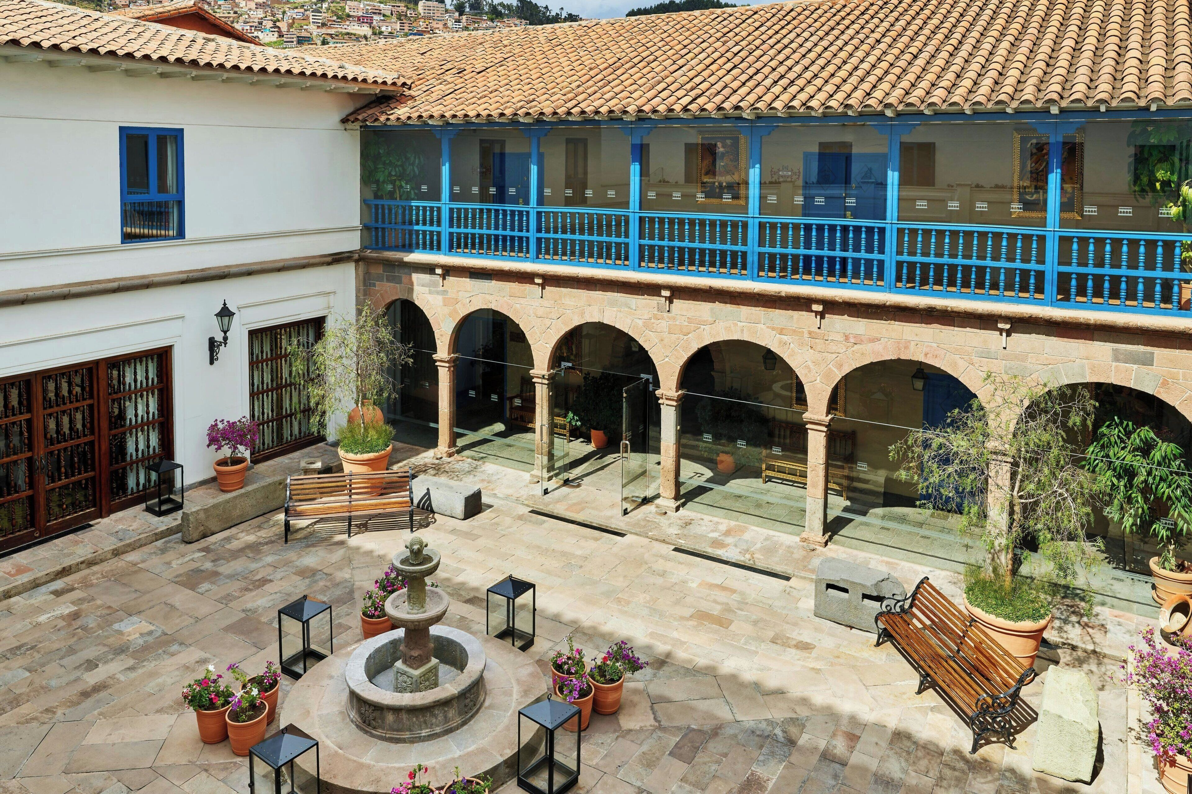 A courtyard with a fountain, benches, potted plants, and a balcony with blue railings.