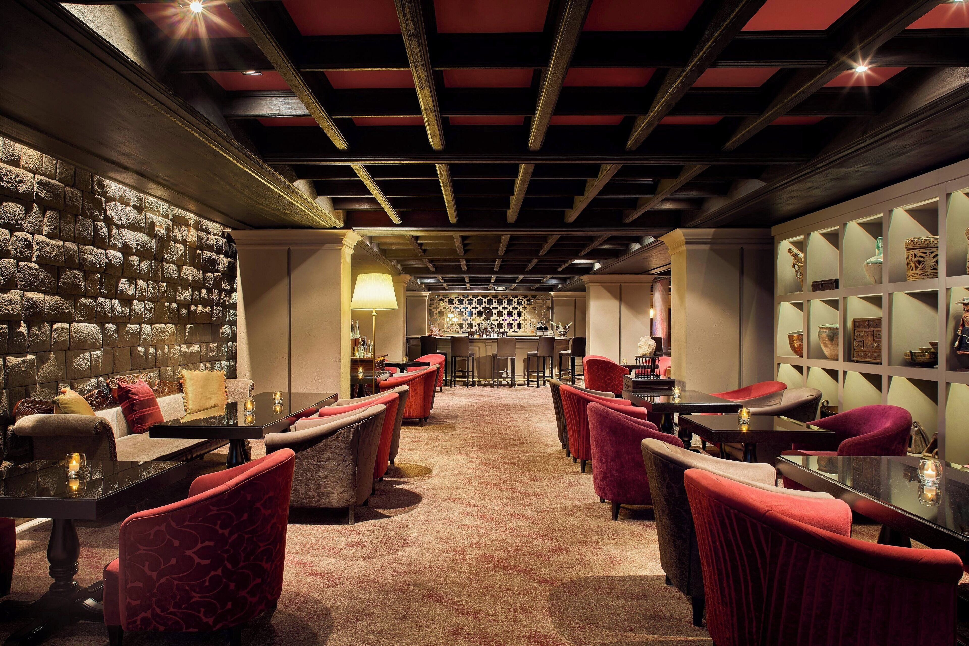 A dimly lit bar area with red upholstered chairs and tables, a stone wall, and a bar counter with stools.