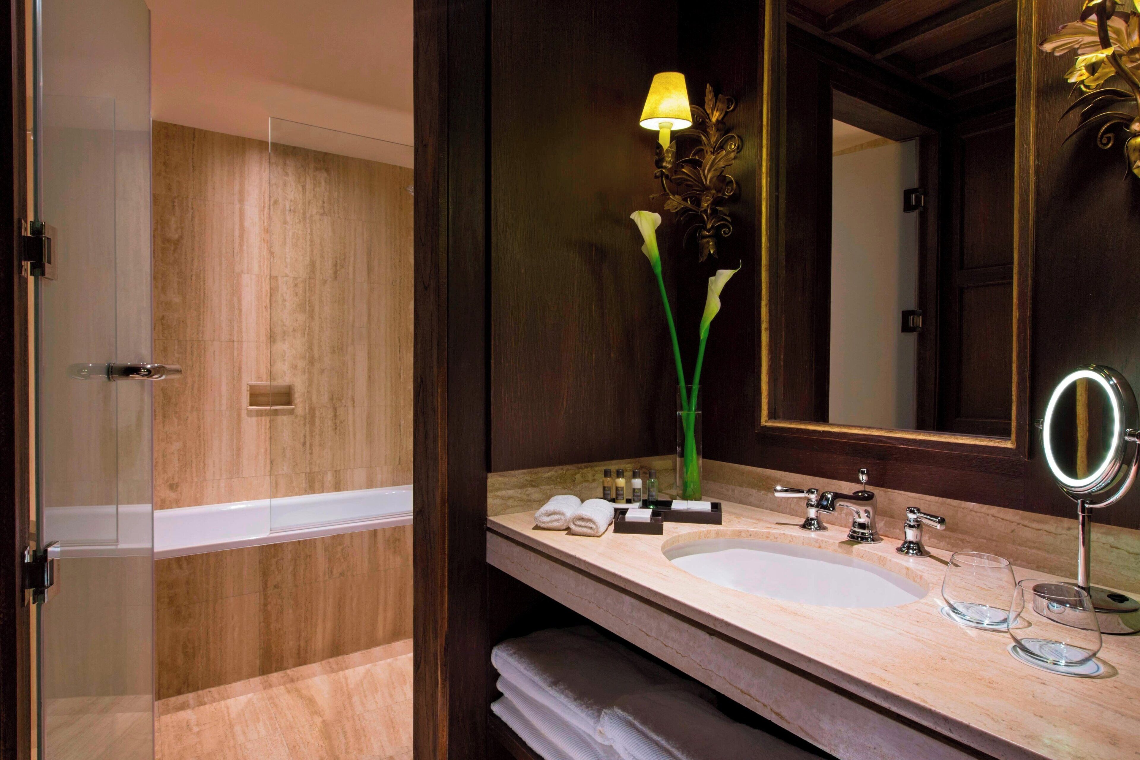 A bathroom with a marble sink, two faucets, a mirror with a round frame, a vase with flowers, and a glass shower enclosure.