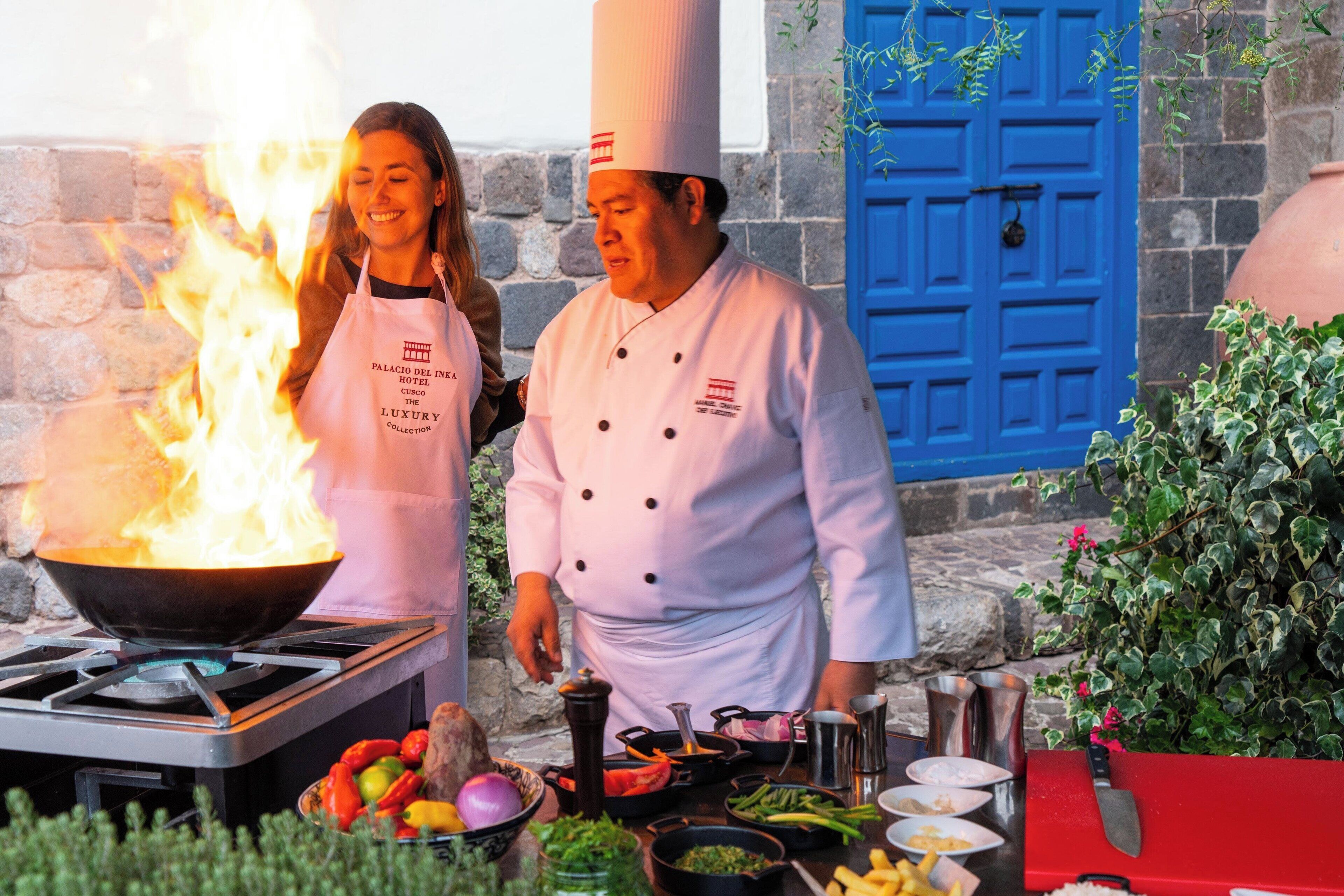 A chef preparing food outdoors with a large fire, a blue door, and stone walls in the background.