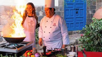 A chef preparing food outdoors with a large fire, a blue door, and stone walls in the background.