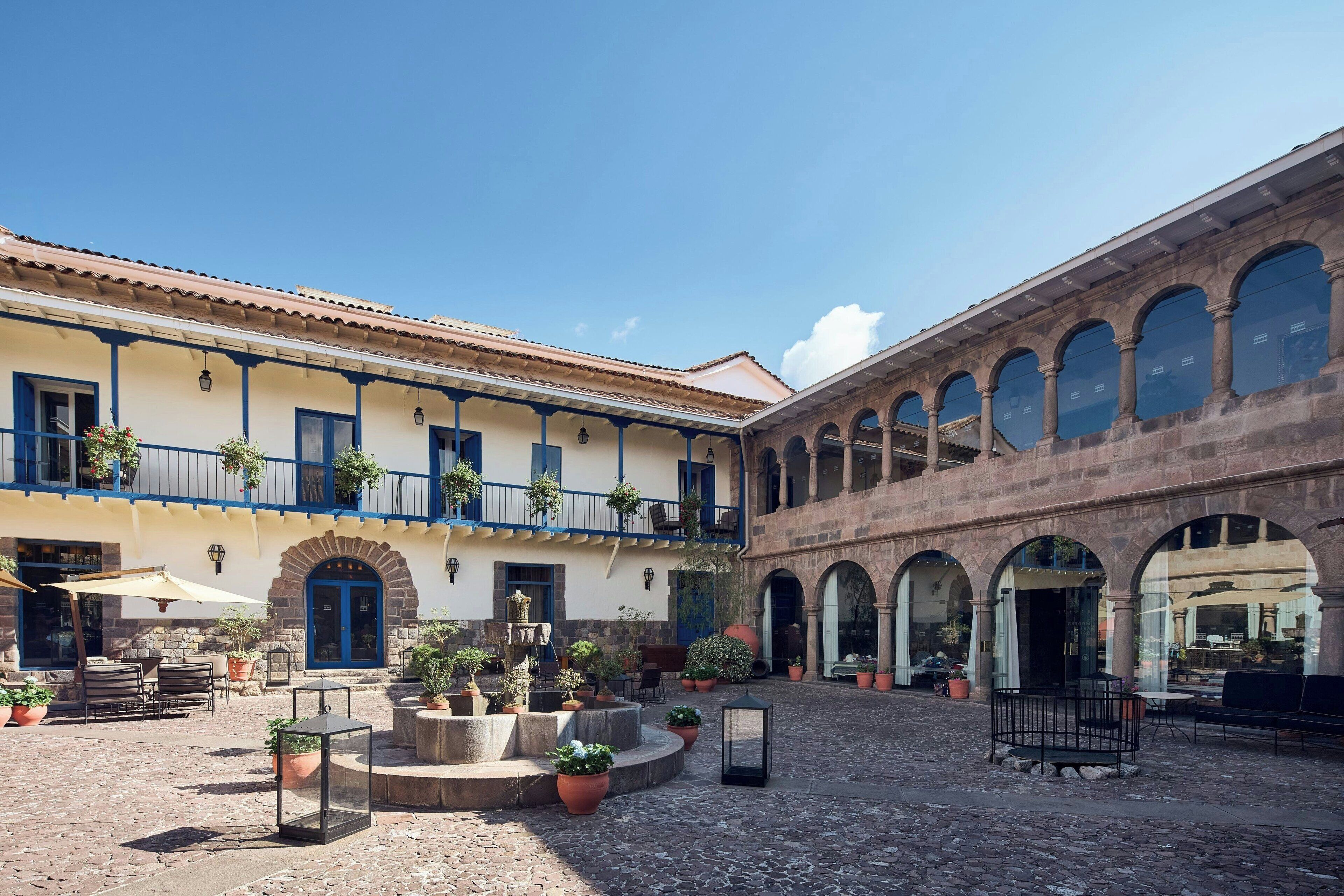 Un patio con fuente, plantas en macetas, asientos exteriores y un edificio de piedra con arcos.