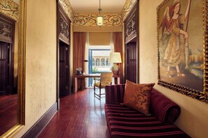 A hallway with a wooden floor, a sofa with a patterned cushion, a chair, and a table with a lamp.