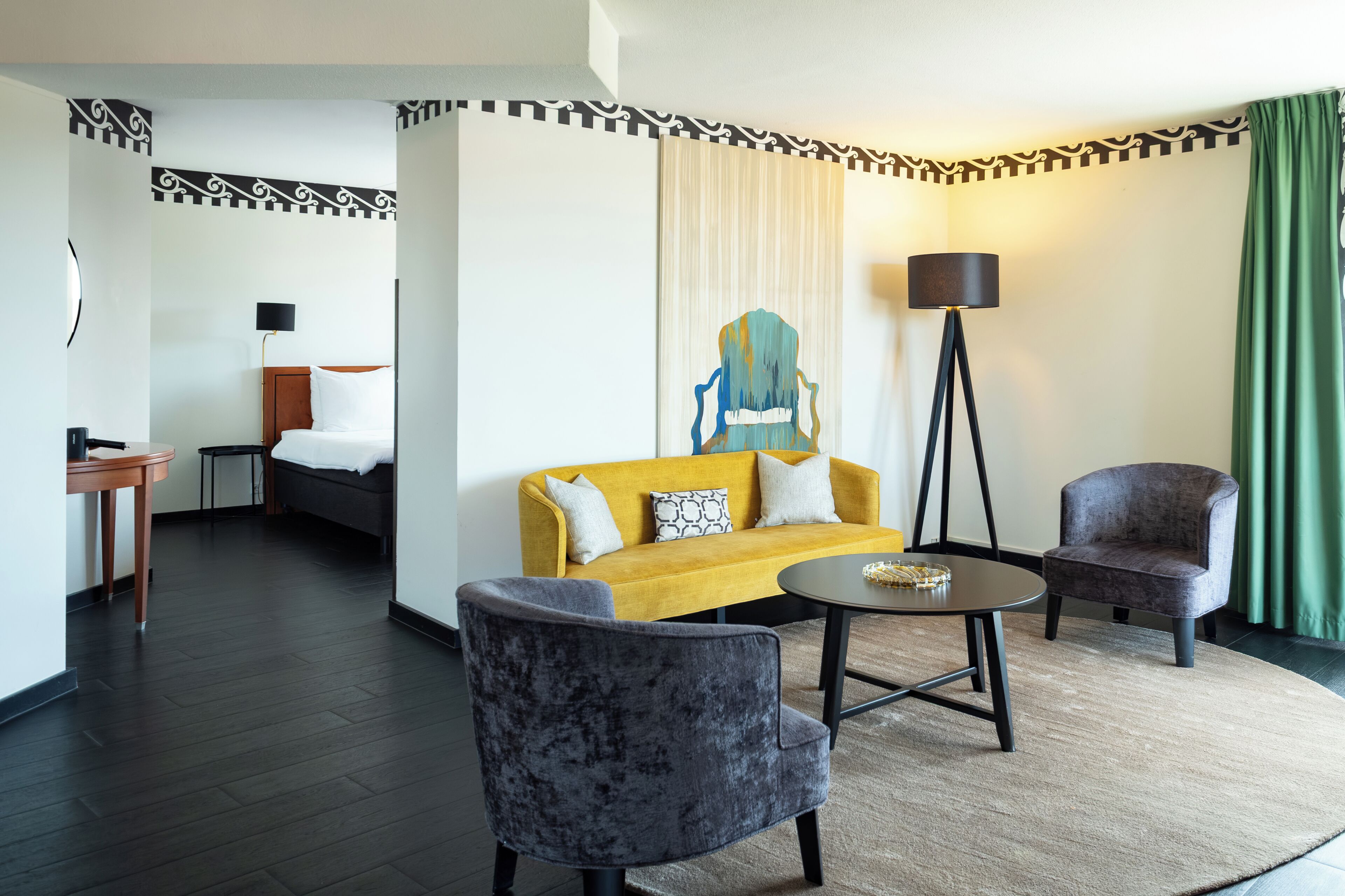 Een hotelkamer met een bed, een gele bank, twee fauteuils, een kleine tafel en een vloerlamp.